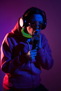 Cyberpunk woman in hooded hoodie and sunglasses dances against  wall with neon sticks and headphones