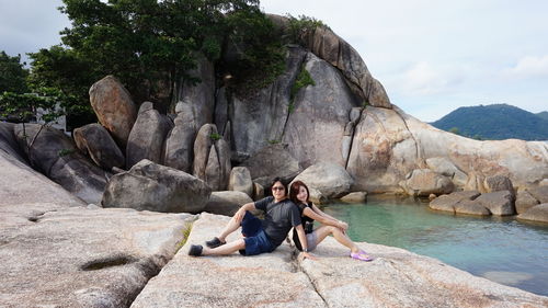 Portrait of couple sitting on rock formation at beach against sky