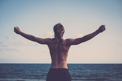 Rear view of shirtless man standing with arms outstretched by sea against sky