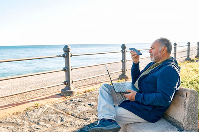 Handsome bearded mature man with laptop working outdoors while sitting on bench at the seaside. 