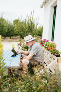 Elderly man in casual clothes and hat sitting on wooden bench at shabby table and browsing data on tablet during break in garden outside house on summer day in countryside
