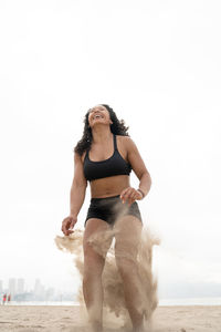 Low angle of cheerful asian female athlete in moment of jumping above sandy seashore during fitness workout in summer