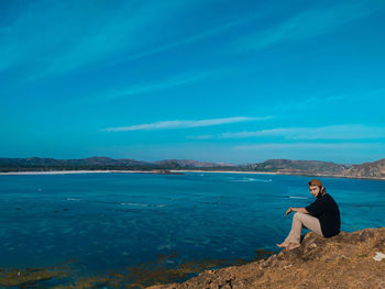 Woman sitting on rocks by sea against blue sky