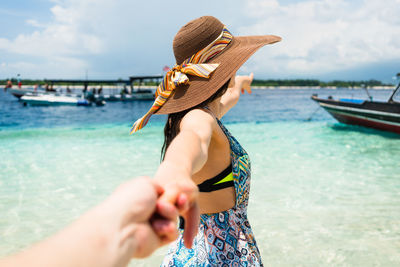 Woman wearing hat holding cropped hand of person while standing on shore against sky