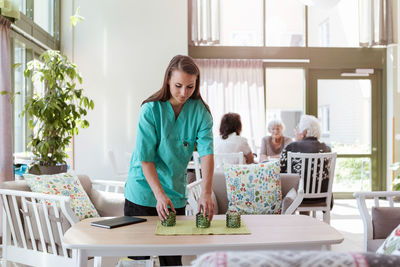 Female caretaker arranging table with senior people in background at nursing home