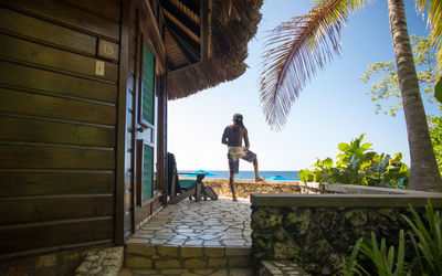 Man standing by a tropical thatched roof beach house on rocky cliff with palm trees in the caribbean