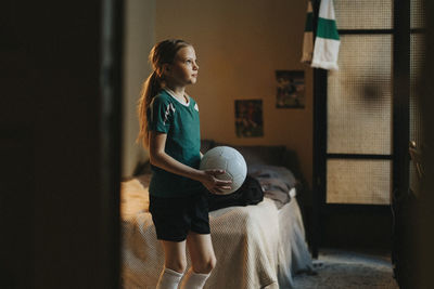 Determinant female athlete with soccer ball standing in bedroom at home