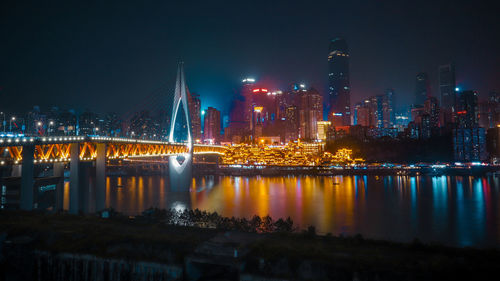 Illuminated bridge over river by buildings against sky at night