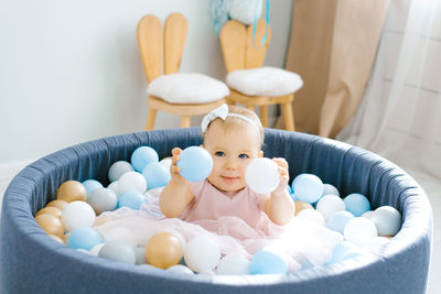 A cute one-year-old baby in a pink dress is bathing in a toy pool with colorful balloons