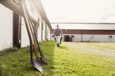 Equipment on grassy field by barn while farmer walking with wheelbarrow