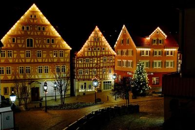 Illuminated street by buildings against sky at night