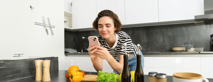 Portrait of young woman using mobile phone while standing in kitchen