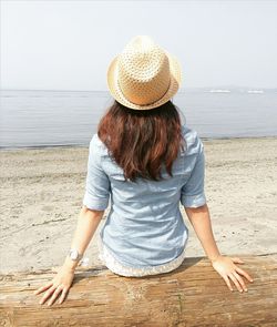 Rear view of woman sitting on driftwood at beach against sky