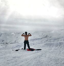 Portrait of shirtless man flexing muscles while standing on snowcapped mountains against cloudy sky