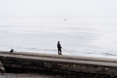 Rear view of man fishing on pier by sea