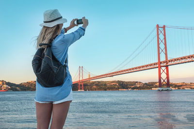 Young traveler woman with smartphone takes pictures in lisbon city near bridge in sunny summer day.