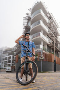 Full body of latin male in protective helmet with photo camera looking up while sitting on bicycle near residential building on street of city