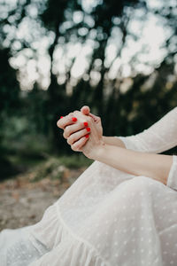Unrecognizable female with red manicure wearing long white dress sitting on ground in forest with green trees on blurred background