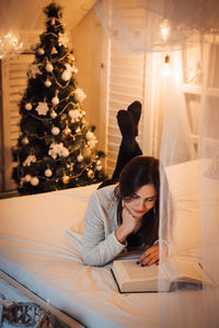 Woman reading book while lying on bed at home during christmas