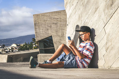 Full length of boy using phone while sitting outside building on sunny day