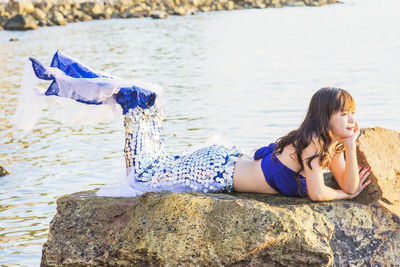 Smiling woman in mermaid costume relaxing on rock in lake