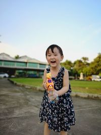 Portrait of cheerful girl holding toy while standing on footpath against sky