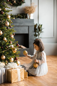 Happy little girl 5-6 year old holding christmas balls decorating tree in room. 