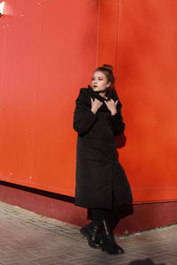Fashionable teenage girl wearing black warm clothing standing against red wall during sunny day