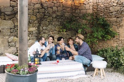 Happy friends with wine partying in yard against stone wall during sunset
