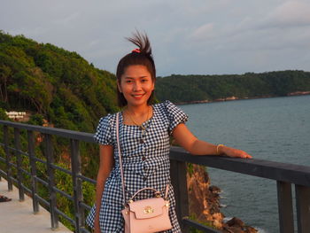 Portrait of smiling young woman standing by railing against sky