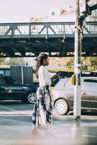 Side view of young woman using mobile phone while standing on sidewalk in city