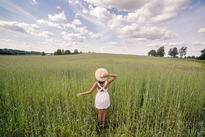 Young woman with long dark hair, on green field with tall grass in a short white dress and straw hat