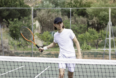 Long-haired young man with a cap playing a tennis match on a court. trying to hit the ball