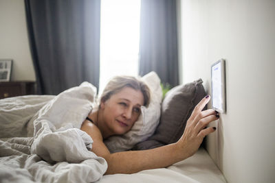 Woman using digital tablet mounted on wall while lying on bed at smart home