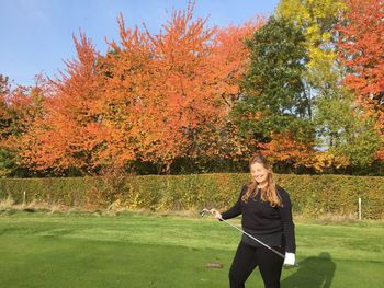 Young woman holding golf club while standing on golf course against autumn trees