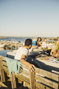 Rear view of man with friends sitting in background at harbor against clear sky