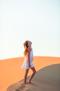 Side view of girl standing at desert against clear sky during sunny day