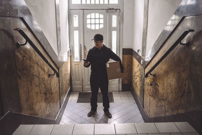 Delivery man using phone while holding package against door