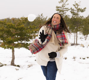 Portrait of happy woman throwing snow while standing against trees