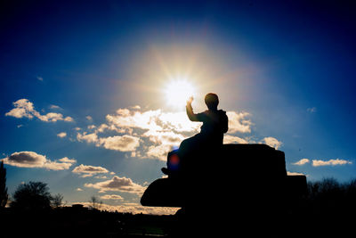 Low angle view of silhouette man gesturing while sitting on rock against sky