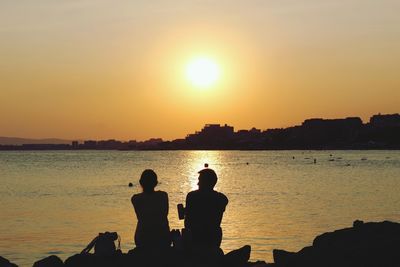 Silhouette people sitting by sea against sky during sunset