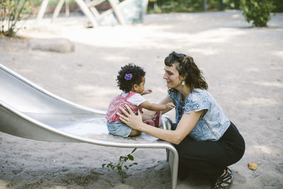 Side view of mother supporting daughter sitting on slide at playground