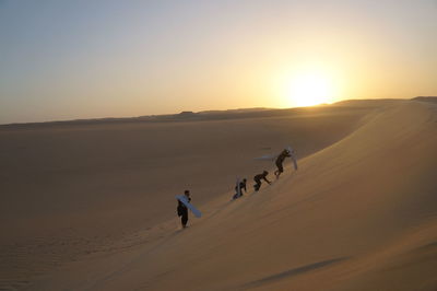 People with surfboards on sand dune at desert against sky during sunset