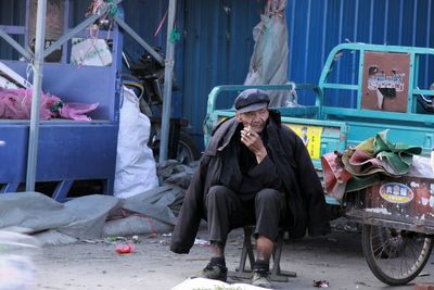 Homeless man smoking cigarette while sitting on stool in city