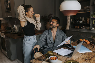 Non-binary person brushing teeth while talking to man sitting at dining table in home