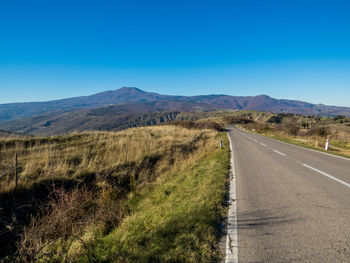 Road leading towards mountains against clear blue sky