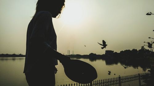 Side view of silhouette woman standing by lake against sky during sunset