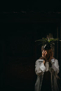 Woman covering face with pineapple standing outdoors at night