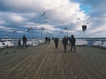 People on boardwalk with seagulls flying against cloudy sky