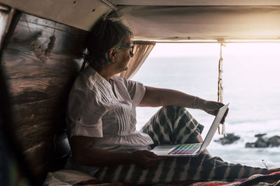 Senior woman sitting in vintage van by he sea, using laptop